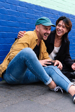 Happy gay couple sitting on sidewalk in front of wall