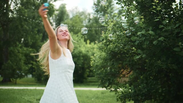 Woman in white dress plays with bubbles in sunny park. She smiles, arms outstretched, enjoying outdoor fun. Bubbles float around her, catching light. Ideal for summer, celebration