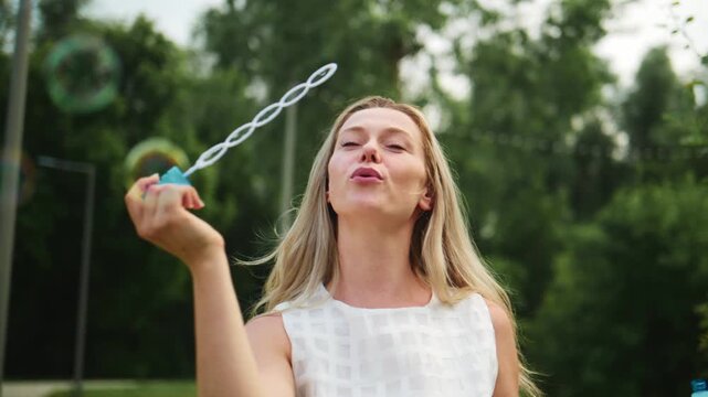 Young woman blows bubbles with joyful expression outdoors. Her blonde hair flows freely as she smiles. Bubbles float gracefully through green trees. Footage ideal for summer, childhood