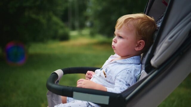 Cute toddler in stroller reaches out while watching bubbles float by. Soft focus captures serene outdoor moment. Child shows curiosity and wonder in green park setting. Ideal for parenting, childhood