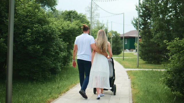 Couple walks together with stroller on tree-lined path. Back view shows affectionate hold and relaxed stroll. Sunlight filters through greenery, highlighting natural calm. Ideal for family, parenting