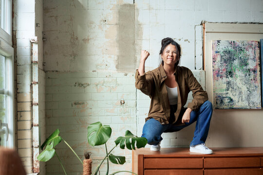 Excited painter showing clenched fist squatting on cabinet by paintings at art studio