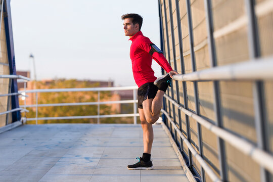 Jogger with smartphone in arm pocket, stretching his leg on a bridge railing