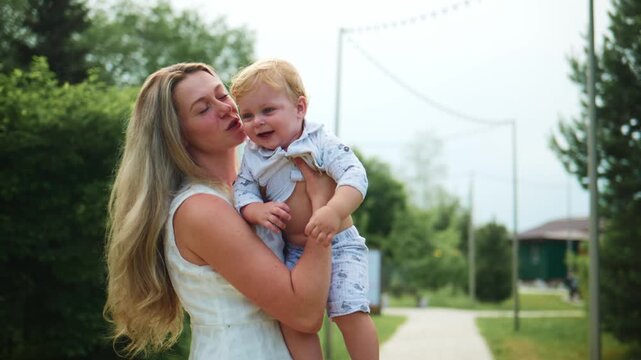 Mother lifts toddler in joyful outdoor moment. Child laughs, arms outstretched. Mother smiles warmly, eyes bright with delight. Scene captures playful, carefree family bonding
