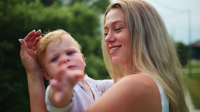 Mother holds baby gently outdoors. Mother smiles softly, eyes focused on child. Child waves hand with curious expression. Scene captures tender bond between parent and child