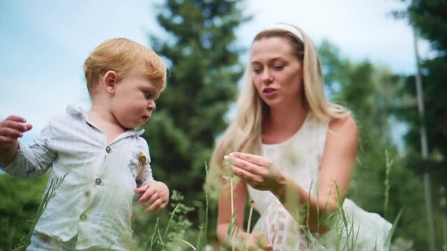 Mother and toddler explore tall grass together. Mother smiles warmly, guiding child's hands. Child focuses intently on blades of grass. Scene captures gentle, playful outdoor bonding
