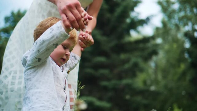 Mother gently holds her child's hands while walking through a sunlit garden. The child looks down, focused on the grass beneath them. Tall green trees and soft wildflowers surround them