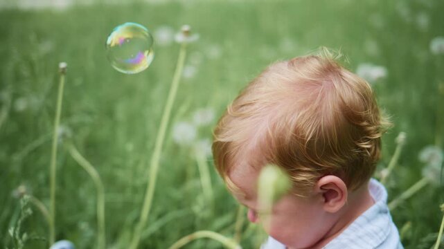 Young child watches bubble float near grass. Child's face shows curiosity. Gentle outdoor moment. Ideal for nature or childhood themes. Soft focus enhances dreamy atmosphere