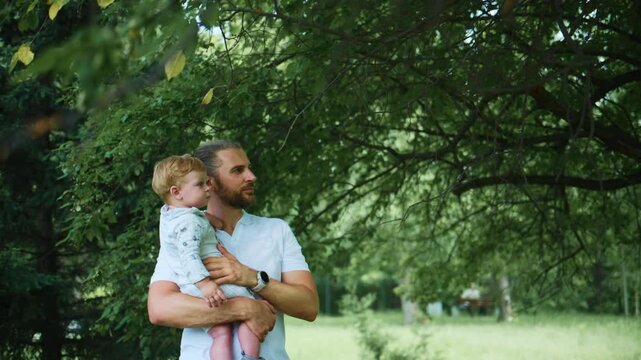 Father holds toddler under leafy tree. Child points upward with curiosity. Man smiles, enjoying outdoor moment. Green foliage frames family interaction. Ideal for parenting, nature