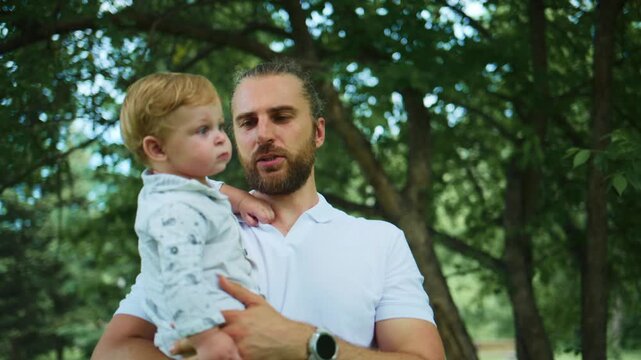 Father holds his young child in a forest. The child points upward with curiosity. The father smiles softly, enjoying the moment. Nature surrounds them with green trees and dappled sunlight