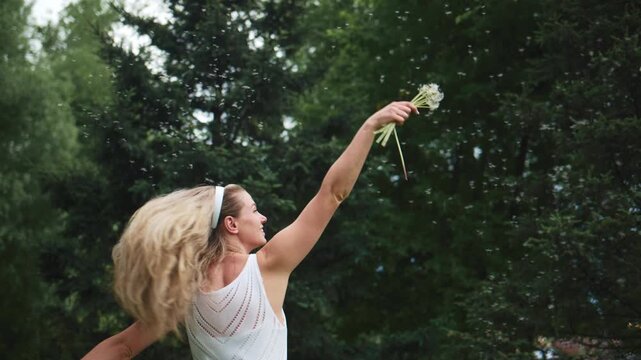 Blonde woman in white dress joyfully tosses flower bouquet upward. She smiles brightly outdoors, surrounded by green trees. Motion blur suggests energetic movement. Ideal for wedding, celebration