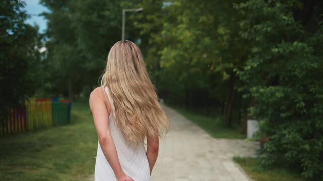 Woman walks down a tree-lined path. She wears a white dress, hair flows freely. The path winds through greenery, sunlight filters through leaves. The scene feels calm, peaceful, and serene