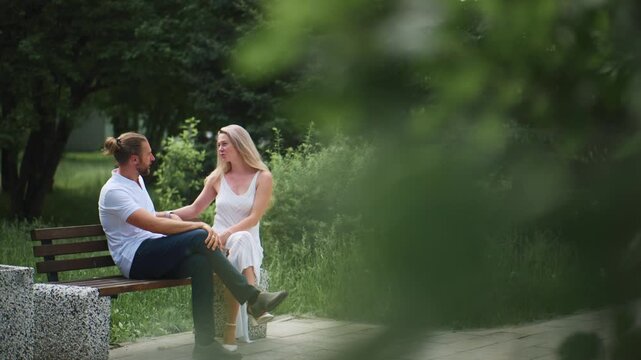 Couple sits close on park bench, smiling and talking. Greenery frames scene naturally. Intimate moment captured through blurred leaves. Ideal for romantic scenes or relationship themes