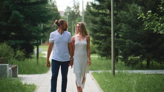 Couple walks hand-in-hand on paved path through lush green park. Man wears white polo, woman wears light dress. Both smile, gaze at each other, appear happy and relaxed. Scene evokes romance