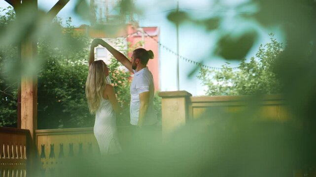 Couple dances joyfully on wooden deck. Green leaves frame the scene naturally. Warm sunlight highlights their happy expressions. Perfect for romantic or summer-themed projects