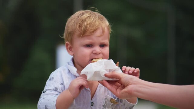 Toddler holds tissue near mouth while adult hand supports. Child smiles brightly, eyes sparkling with joy. Scene captures tender moment of care outdoors. Perfect for parenting, health
