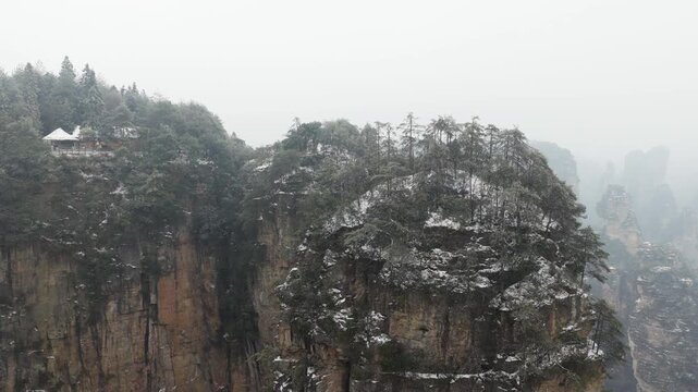 Drone view of Zhangjiajie National Forest Park landscape with towering sandstone pillars and cliffs wearing a fresh layer of snow during winter, trees clinging to the rock formations under a foggy sky