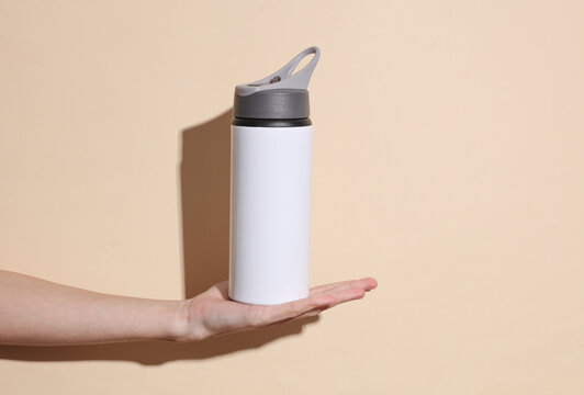 Young woman hand holds white metal flask for water on beige background with shadow