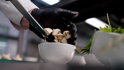 Close up of chef hands in black gloves slicing mushrooms with knife into white bowl. Action moves from slicing to finishing. Professional kitchen setting with green herbs nearby © Ruslan