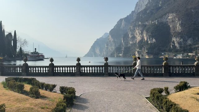 A woman walks a pet dog along Riva del Garda in Italy with Lago di Garda and the surrounding lakeside landscape visible