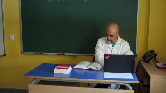 Medium shot of a mature male teacher or healthcare professional wearing a white lab coat sitting at his desk. He types on a laptop computer alongside open reference books.