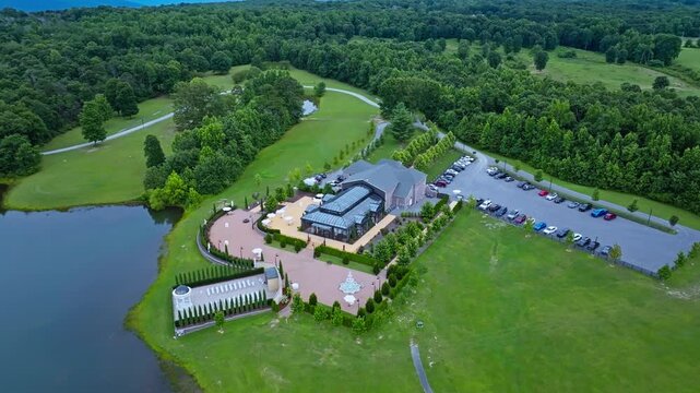 Elegant wedding venue with glass atrium and curved stone patio sits beside calm pond near Trenton, Georgia, crowded parking lot borders dense green summer forestland. Aerial drone top down.