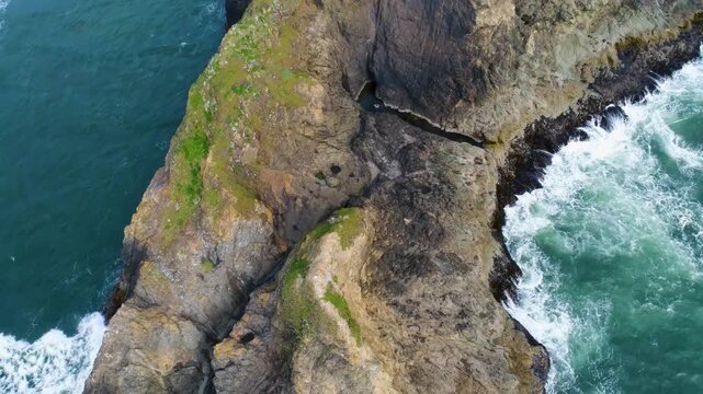 A cinematic drone shot reversing over a sea stack, facing down at the rough Pacific surf crashing into its rugged cliffs, near Ariya&rsquo;s Beach and Branson Carlton&rsquo;s View on Oregon&rsquo;s southern coast.