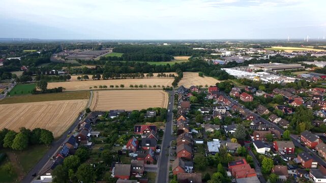 Golden harvested wheat fields border dense red rooftop village in Germany, tall tree rows and distant wind turbines rise beside industrial park amid open countryside. Aerial drone flyover shot