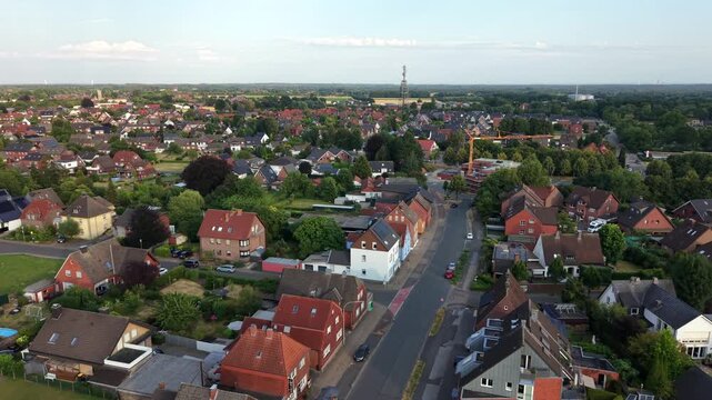 Cars drive along curving street through small German town lined with red brick houses, distant construction crane and radio tower rise above dense rooftops and treetops. Aerial drone view.