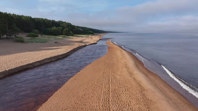 Low altitude drone footage captured at sunrise above the Peterupe River channel flowing alongside a sandy Baltic Sea beach in Saulkrasti, Latvia. Scenic aerial view featuring a narrow sandbar, calm se
