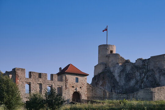 Ancient medieval stone castle ruins with a tower and defensive walls situated on a rocky hill against a clear blue summer sky with a flag.