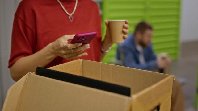 Young adult Caucasian woman using smartphone and holding coffee cup near open cardboard box in storage warehouse. Footage is perfect for e commerce logistics or moving company promotions