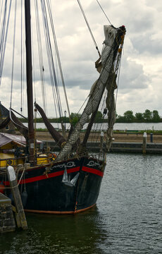 Historic sailing ship docked at a pier, its bow, rigging, and furled sails visible with a cloudy sky above