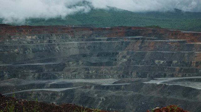 Aerial slow pan across open pit mine with gold, copper ore terraces on mountain slope under low clouds, timelapse.