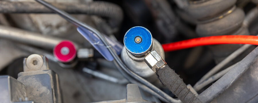 Close-up shows automotive air conditioning service couplers attached beneath open hood during refrigerant refill. Workshop equipment supports vehicle cooling maintenance, diagnostics and repair