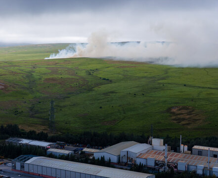Wildfire burning on Llangynidr Moors near Ebbw Vale in South Wales, smoke billowing across dry summer grassland above industrial buildings in valley below