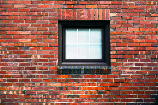 A brick wall with a small square window, blinds drawn.