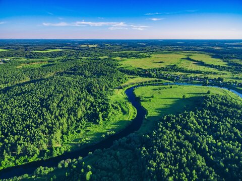 Aerial view of winding river surrounded by lush green forests and open fields in bright daylight in Finland