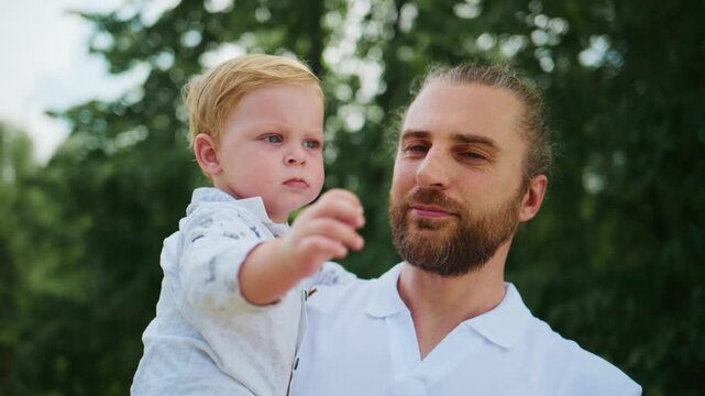 Young father holds baby boy outdoors. Child points ahead with curiosity. Father gazes at child, showing affection. Scene captures joyful family bonding moment. Perfect for parenting, family