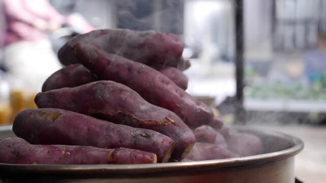 A pile of sweet potatoes steams inside a metal container near a local market street and nearby traffic. Food culture.