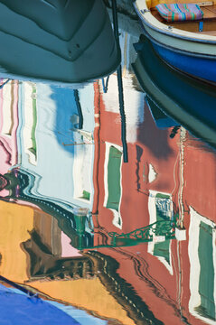 Reflections of red ochre house fronts and boat hulls in the water of the canal at Burano.