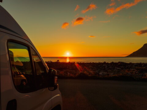 Ramberg, Norway - 11.07.2020: White campervan parked by a rocky shoreline during a vibrant golden sunset over the sea