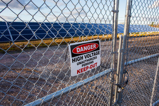A solar farm in the countryside, a secure chainlink fence with a Danger High Voltage sign, saying Keep-Out.