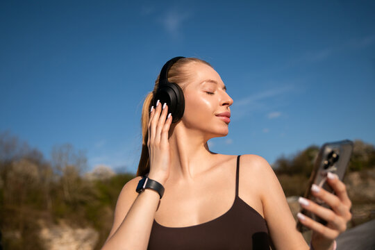 Beautiful woman listening to favorite playlist with relaxed expression. Young woman using smartphone and wireless headphones outdoors. Fitness wellness concept