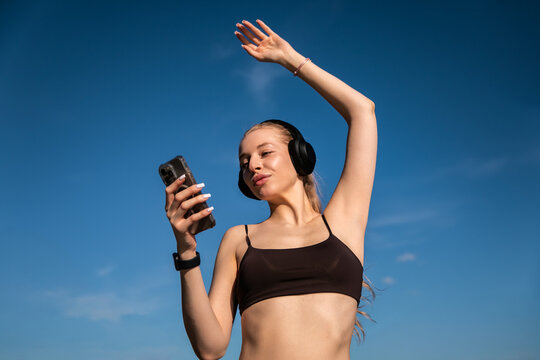 Beauty woman raising arm while listening to wireless music. Attractive woman holding smartphone with joyful expression outdoors. Active lifestyle concept.