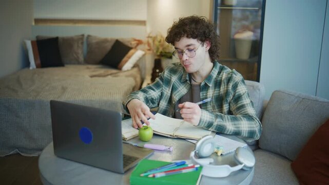 A young man studies at a desk with books and a laptop, taking notes in a notebook before grabbing an apple for a healthy snack while continuing his work