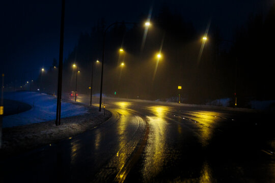 Driver's view of a road merge lane at night, rain on the windscreen, tail lights and tall road lighting reflecting off the west road.