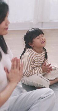 Asian mother and cute daughter sitting on a yoga mat meditating with folded hands together at home.