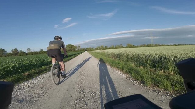 POV first person view from a bicycle riding behind another cyclist on a rural gravel path surrounded by green fields and meadows in Germany under a clear blue sky on a sunny summer day