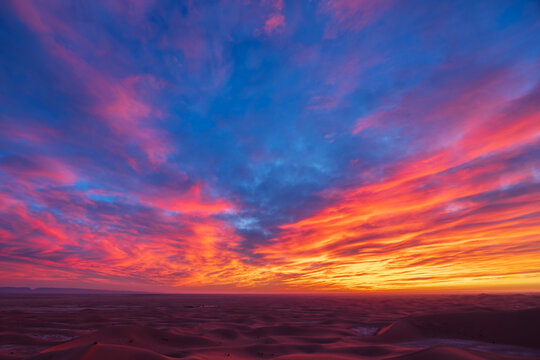 The sky at sunrise above the Chaggaga Sand Dunes, light clouds reflecting red and orange light against blue sky.